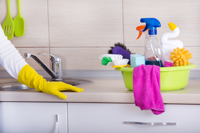 Cleaning supplies in washbasin standing on cleaned kitchen countertop. Hand of cleaning lady with rubber glove beside